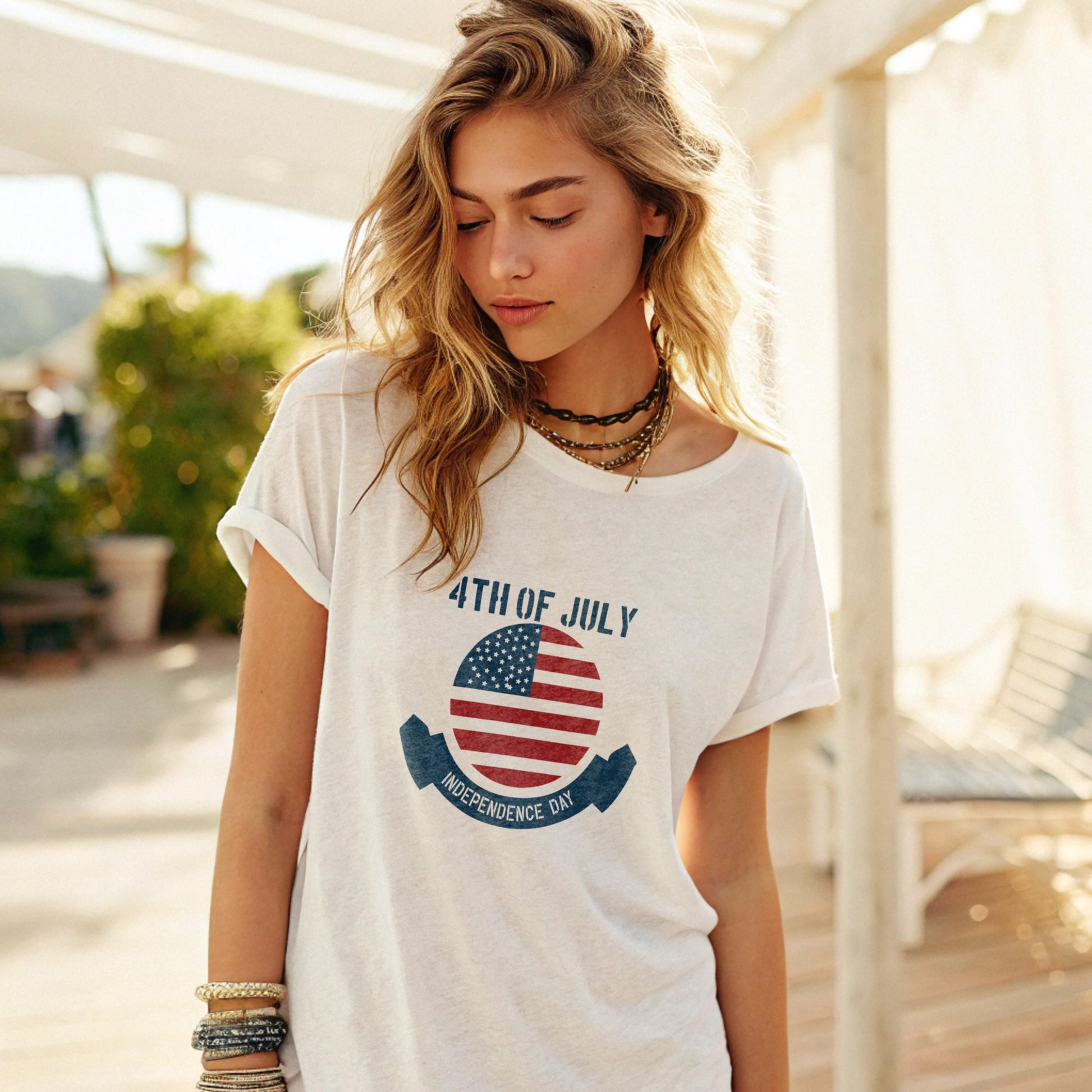 Woman wearing a loose-fitting white T-shirt with a 4th of july print, accessorized with layered necklaces and bracelets, standing on a sunny outdoor patio with greenery and a lounge chair in the background.