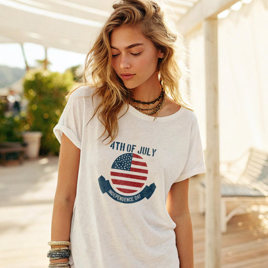 Woman wearing a loose-fitting white T-shirt with a 4th of july print, accessorized with layered necklaces and bracelets, standing on a sunny outdoor patio with greenery and a lounge chair in the background.