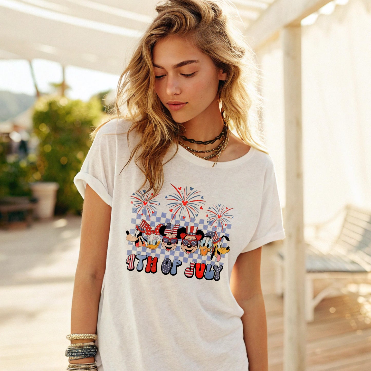 Woman wearing a loose-fitting white T-shirt with a Disney 4th of july print, accessorized with layered necklaces and bracelets, standing on a sunny outdoor patio with greenery and a lounge chair in the background.